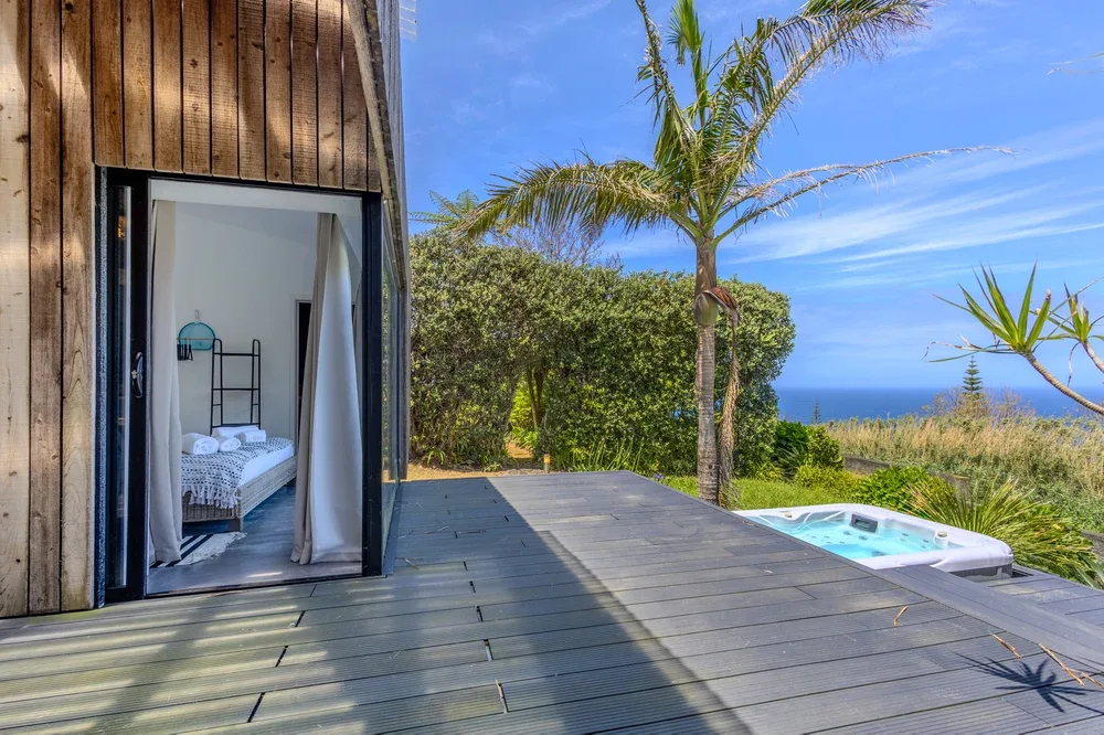 A wooden deck with a hot tub overlooks the ocean beside an open bedroom.