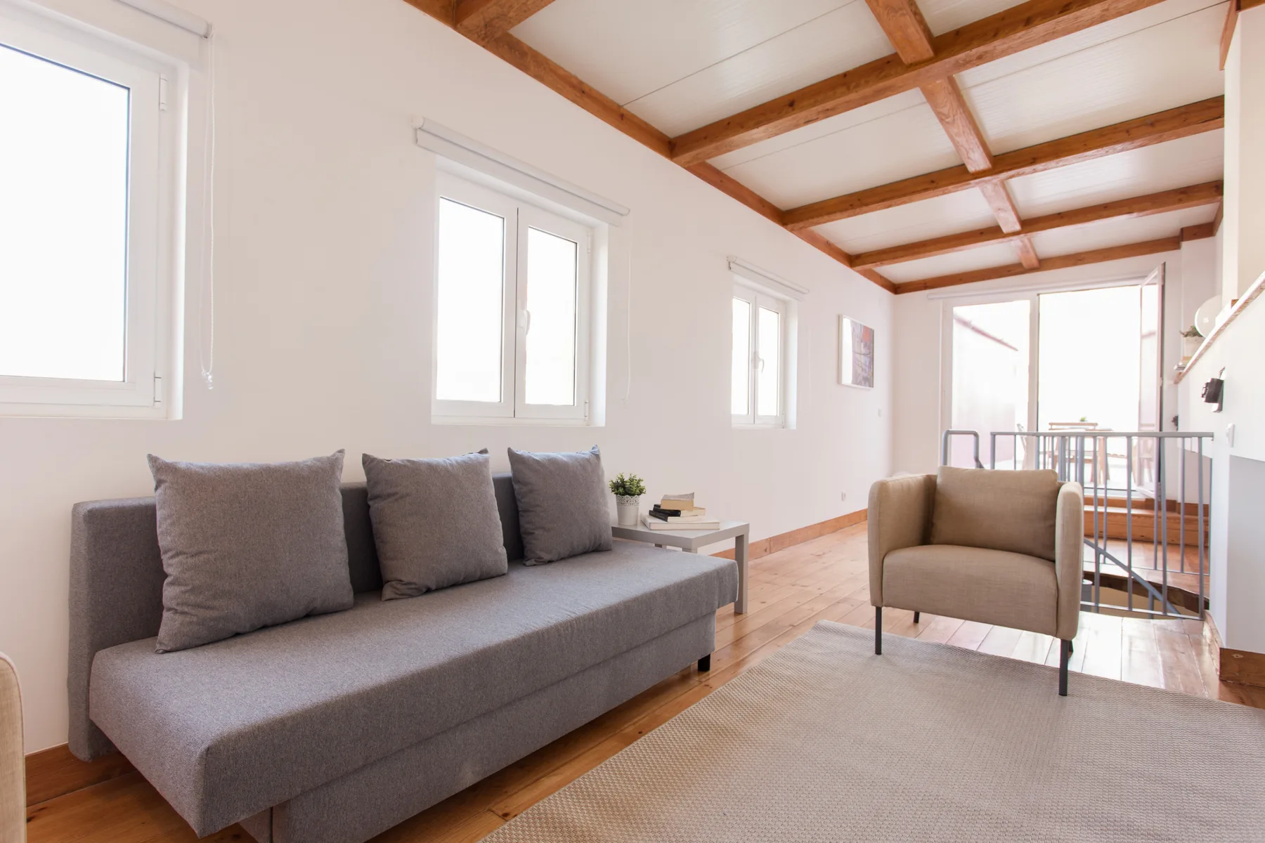 Una sala luminosa con asientos grises, un sillón beige y vigas de madera expuestas en el techo.