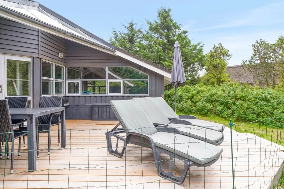 Una terraza de madera junto a una casa tiene asientos reclinables y una sombrilla cerrada.
