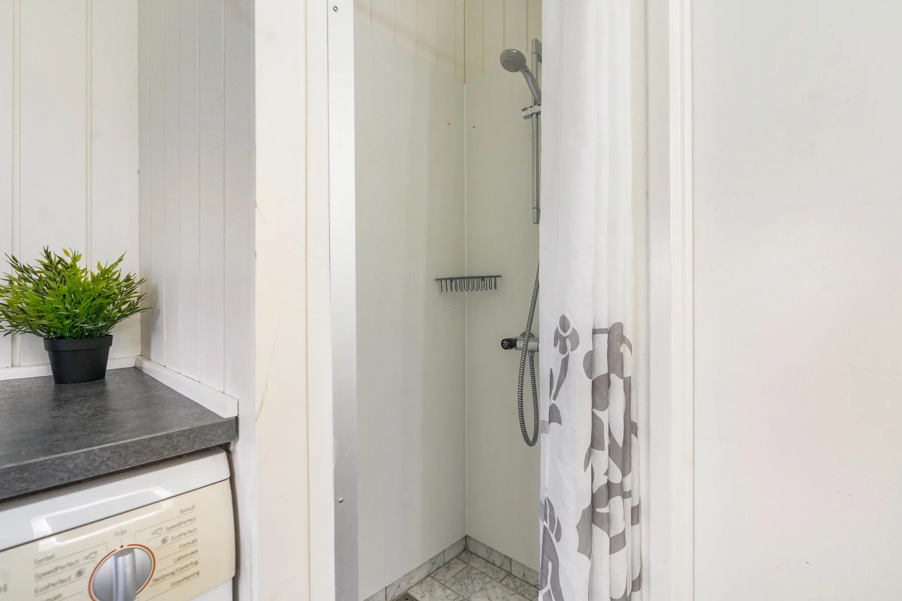 Un baño con ducha y cortina estampada, junto a una planta en la encimera.