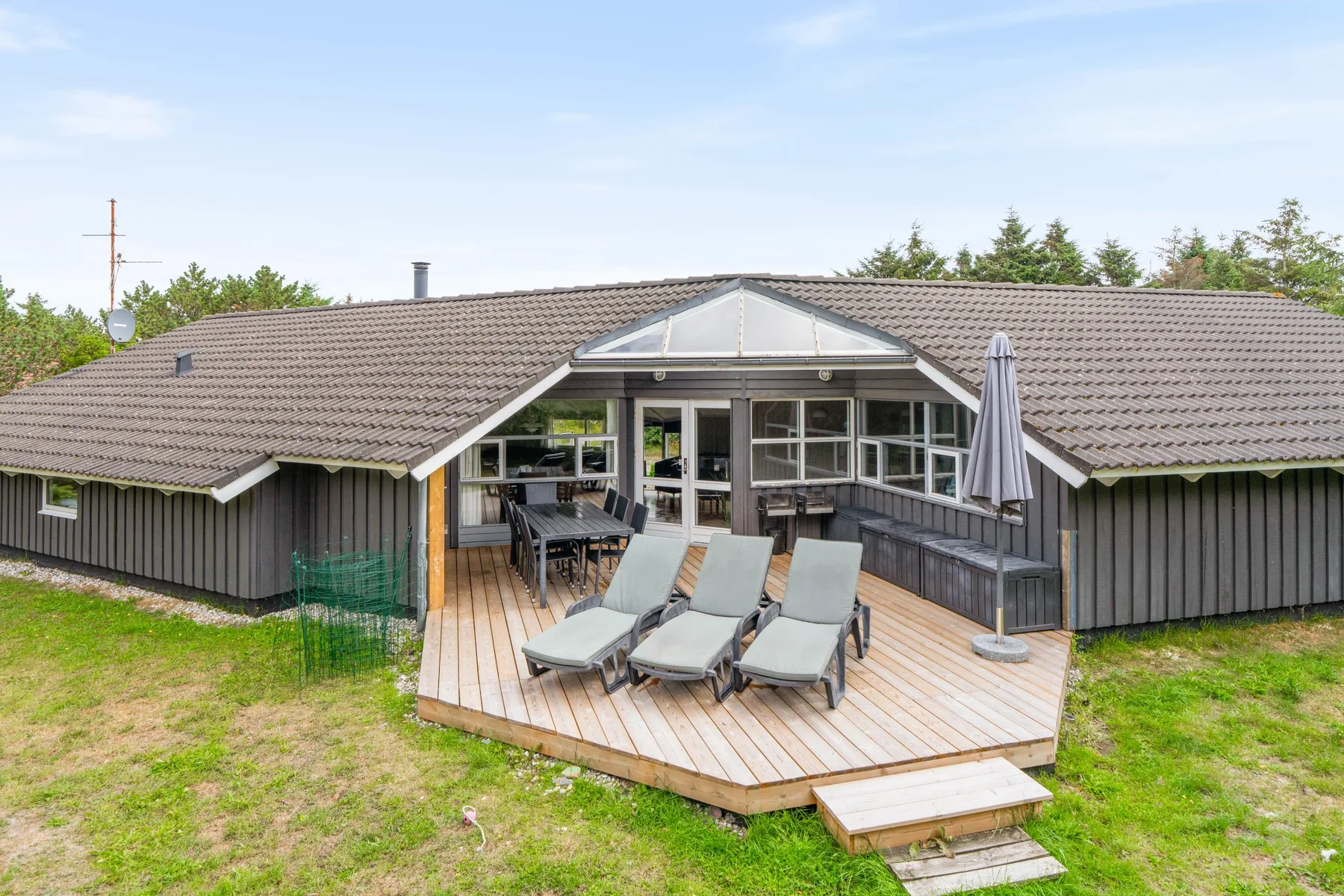 Casa de vacaciones con terraza de madera, asientos al aire libre y una sombrilla plegada.