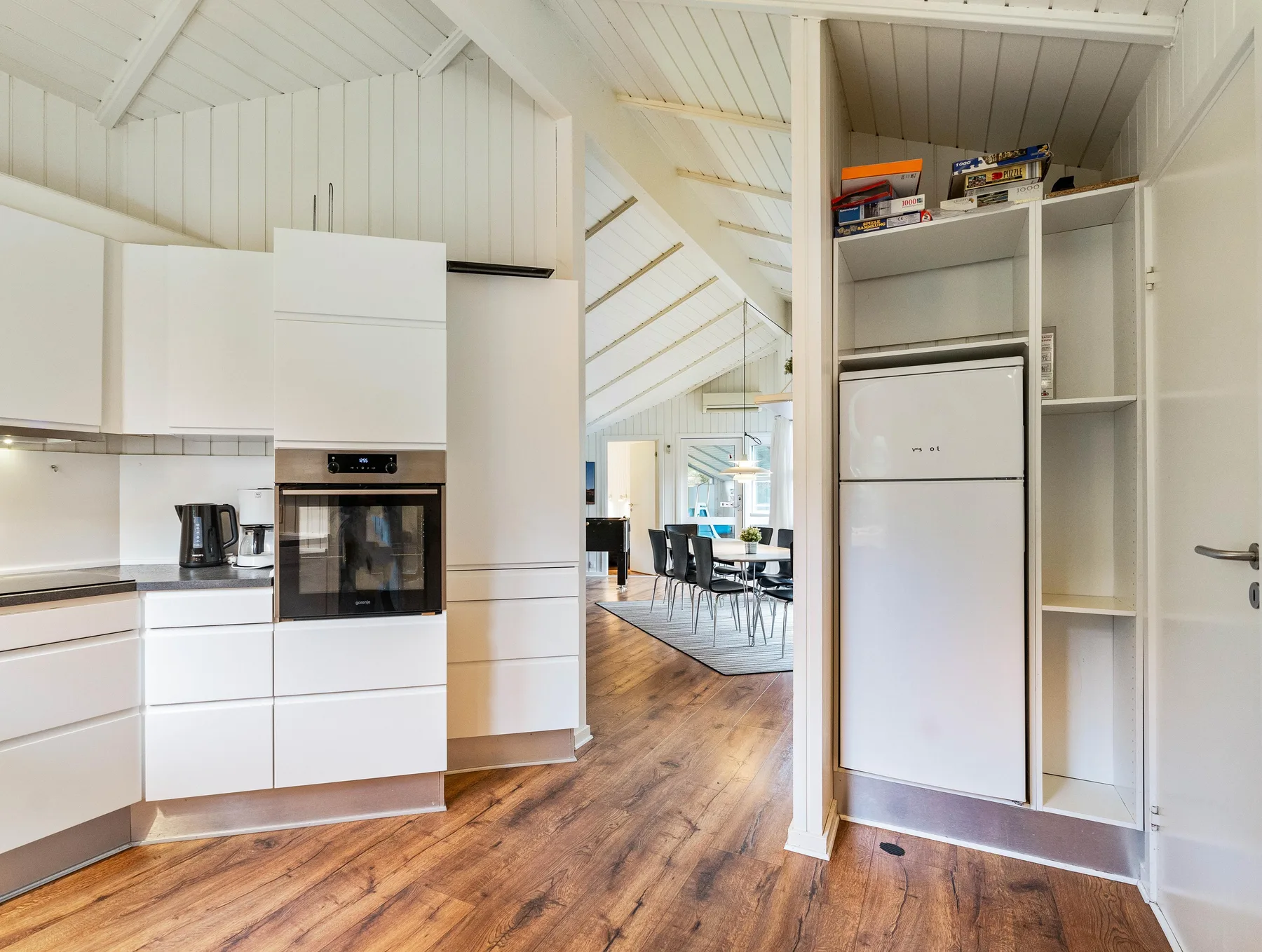 Una cocina luminosa se abre a un comedor, con armarios blancos y suelo de madera.