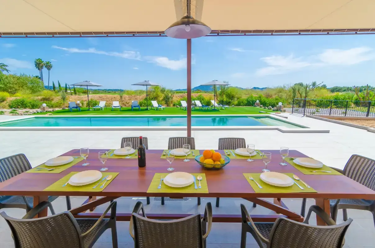 An outdoor dining table is set beside a swimming pool and garden.