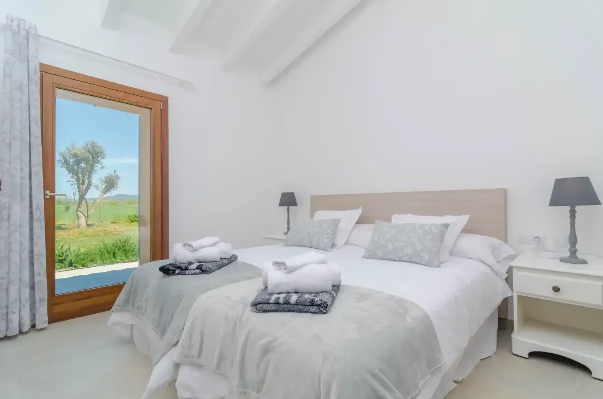 A bright bedroom with a neatly made bed and a glass door opening onto green fields.