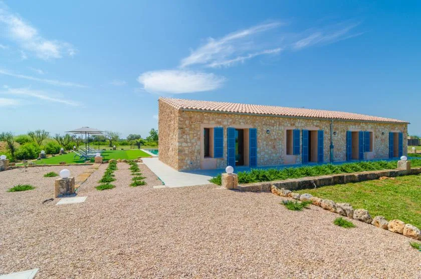A stone building with blue doors stands beside a gravel courtyard and garden.