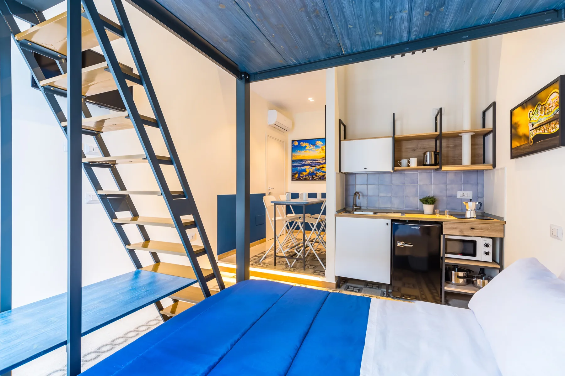 Un espacio compacto para dormir con una cama azul junto a una pequeña cocina y escaleras.
