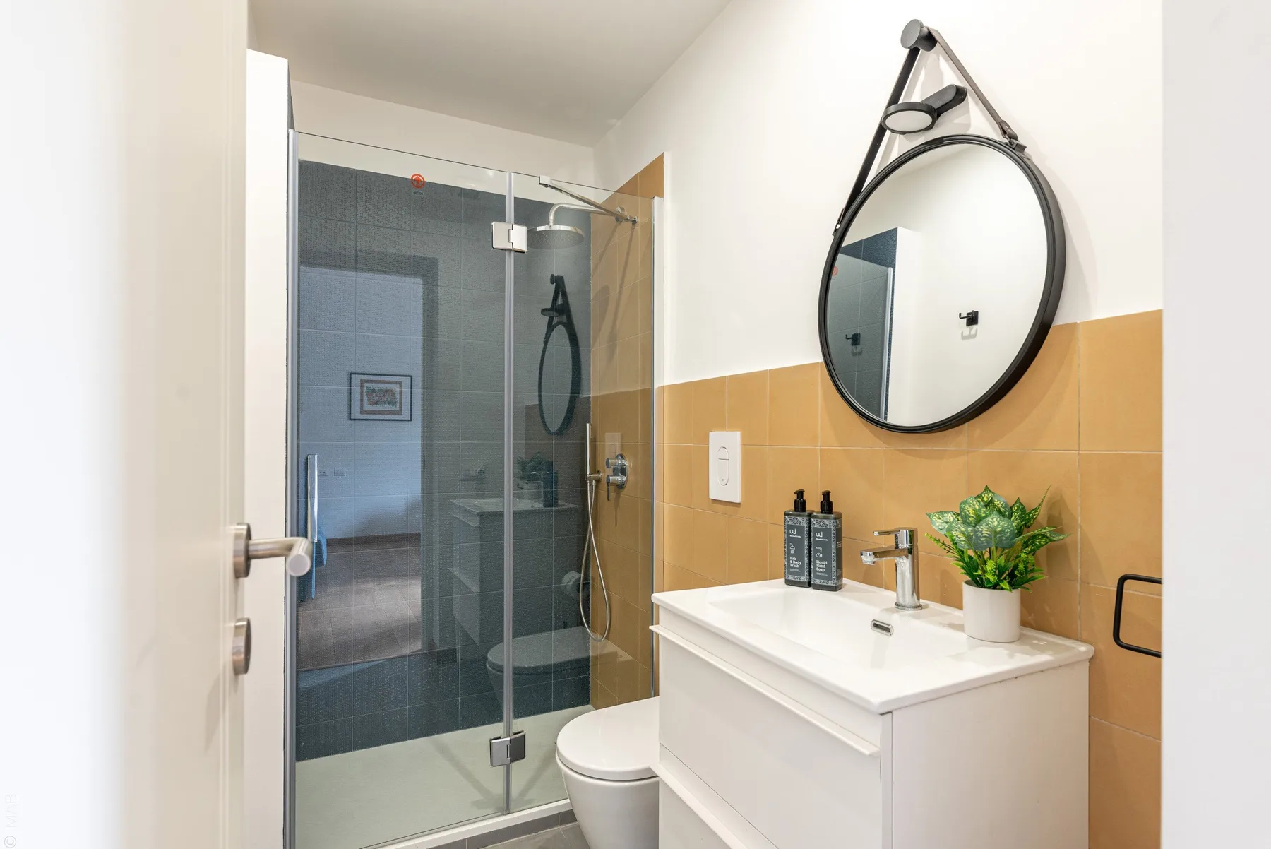 Un baño luminoso con ducha de vidrio, lavabo blanco y espejo redondo.