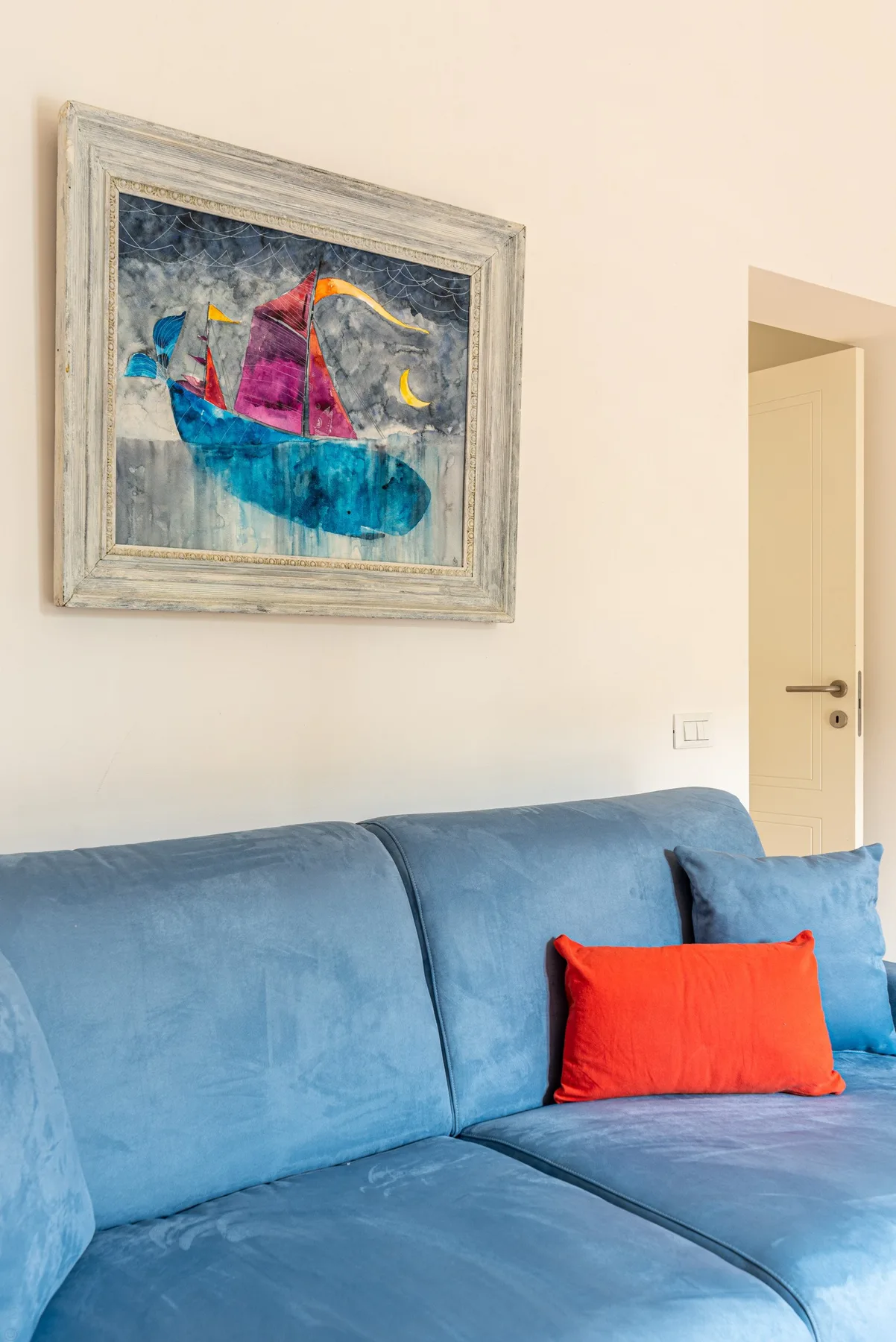 Un sofá azul con un cojín rojo intenso está bajo una pintura enmarcada.
