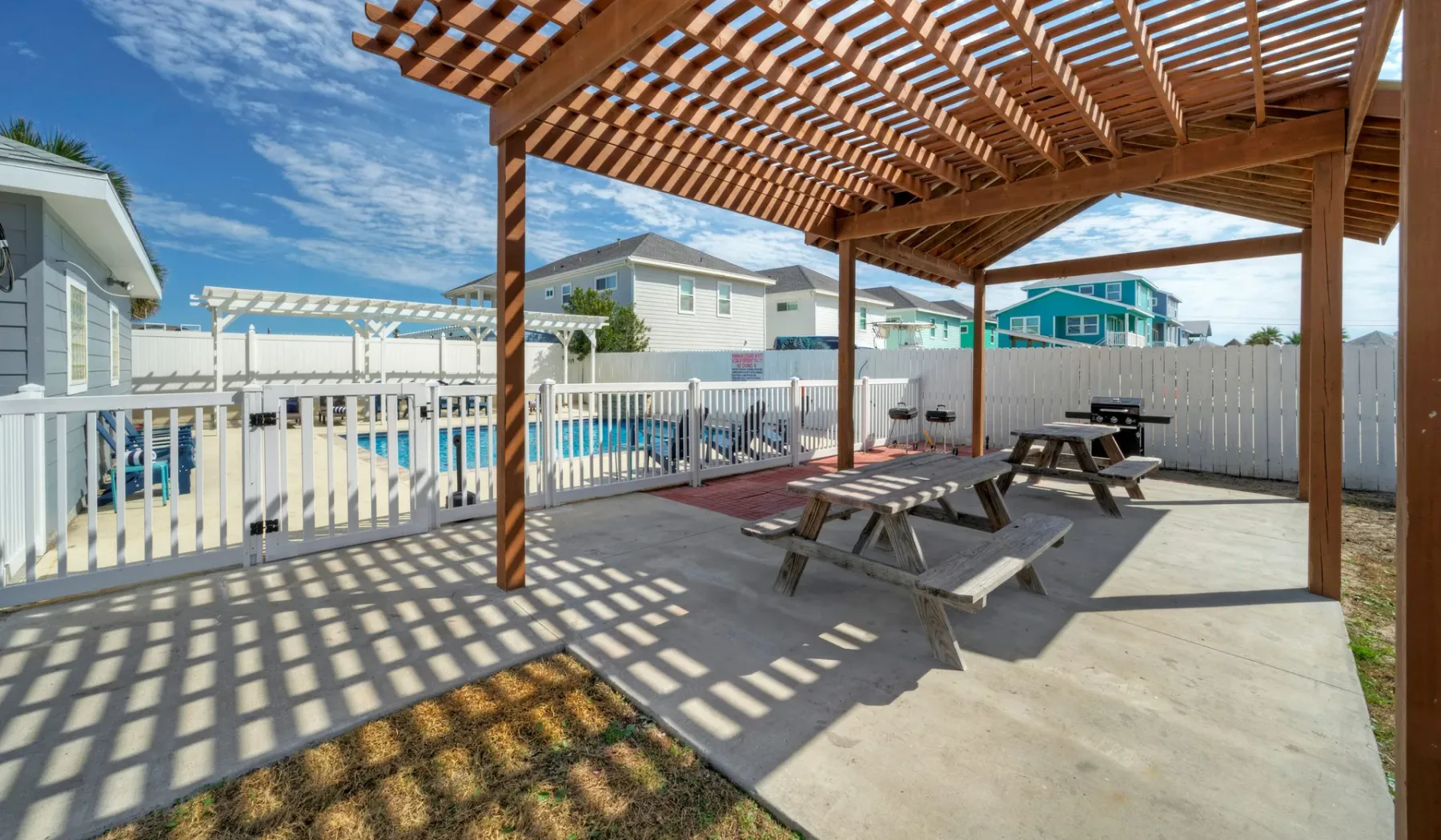 Patio exterior cubierto por una pérgola, con mesas de picnic junto a una piscina cercada.