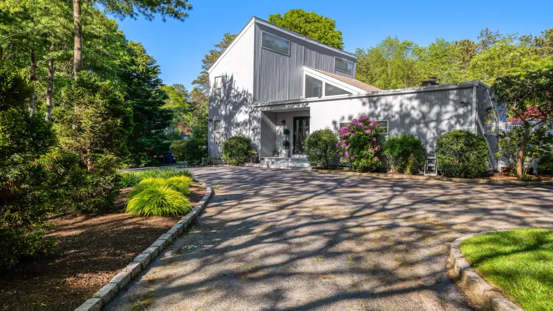 A modern house stands at the end of a paved driveway, surrounded by trees.