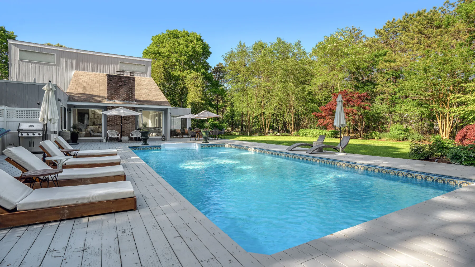 An outdoor swimming pool beside a wooden deck, lounge seating, and a modern house.