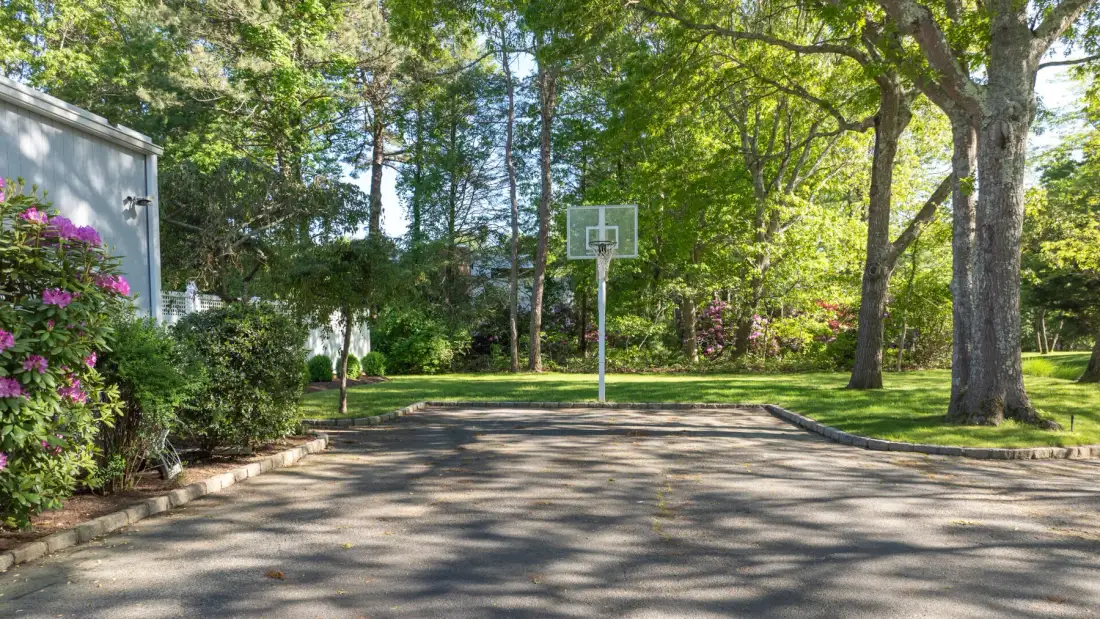 An outdoor basketball court sits beneath leafy trees.