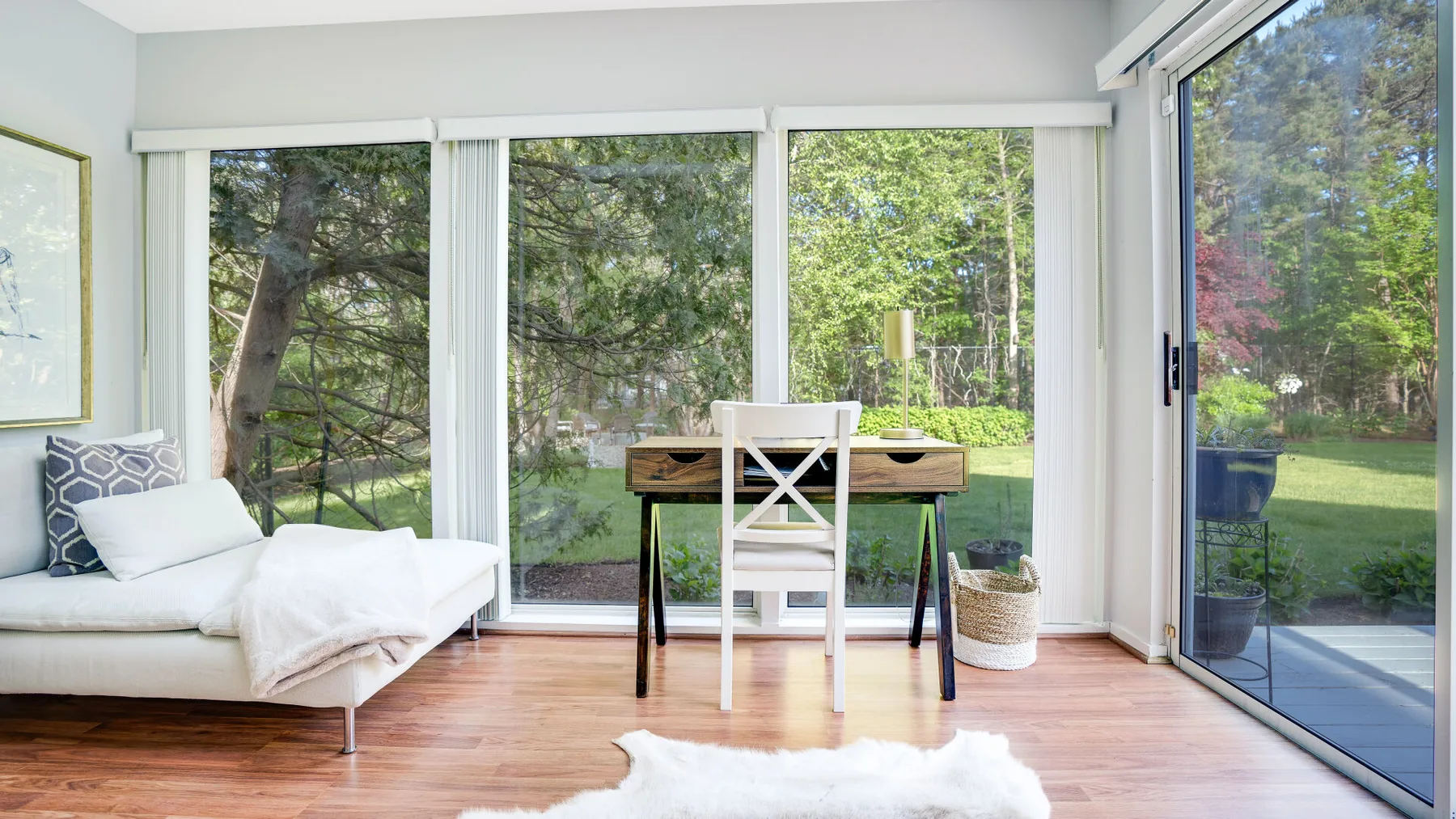 A bright sitting area with a white sofa, desk, and large windows overlooking greenery.