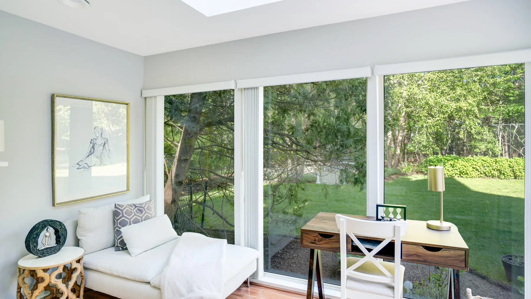 A bright room with a desk and seating beside large windows overlooking a garden.