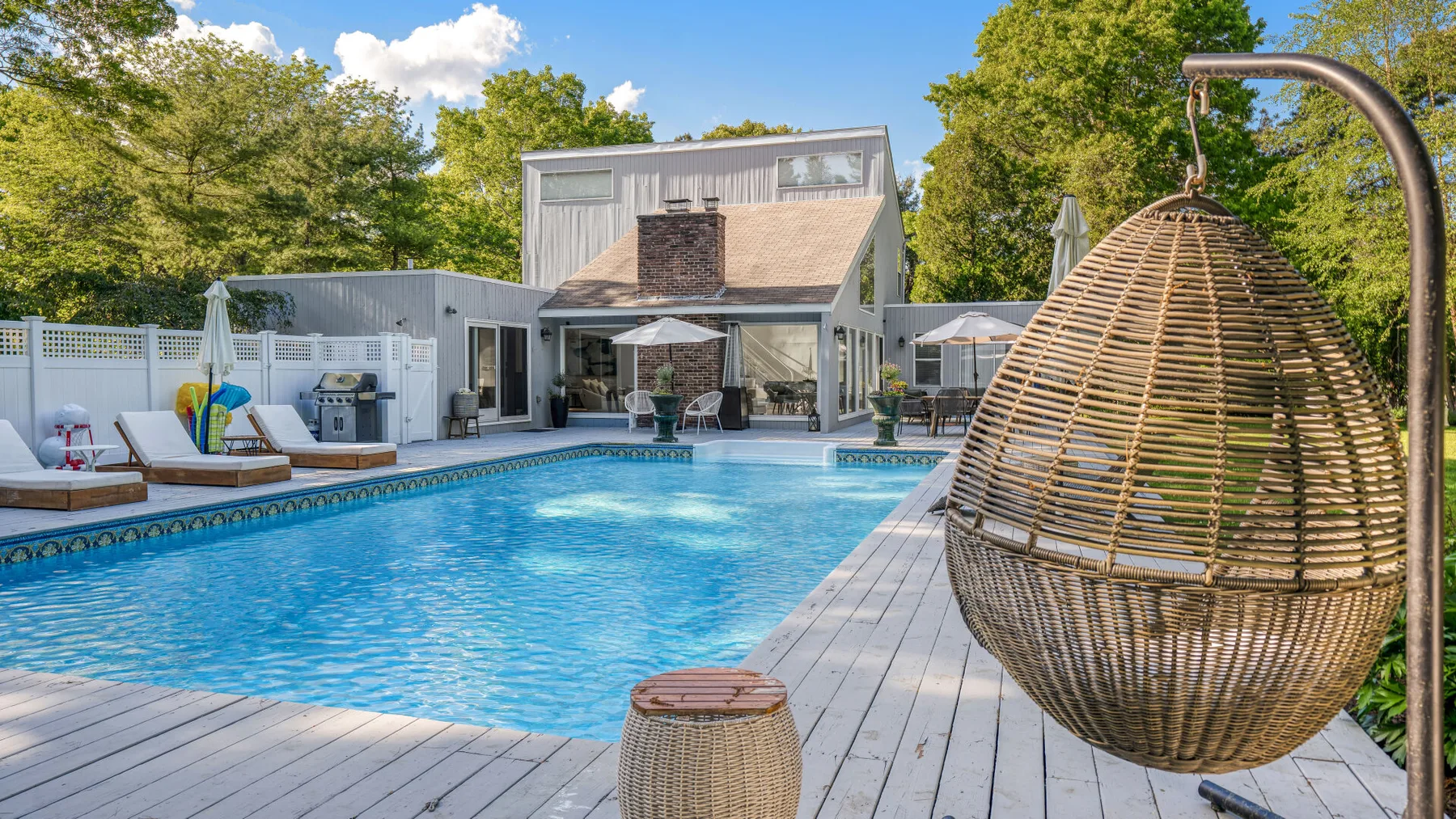 An outdoor pool beside a deck, with a hanging wicker seat and trees.