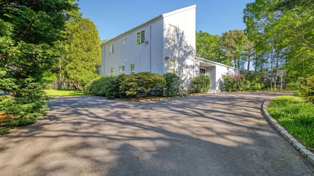 A white building stands beside a paved driveway surrounded by trees.
