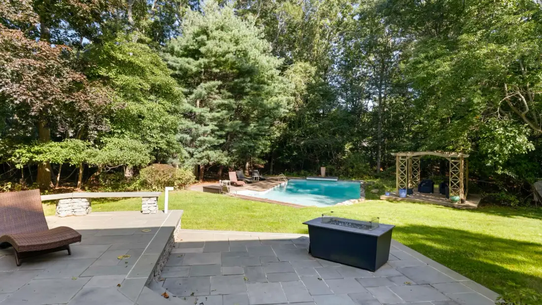 A backyard patio with a pool and a lawn surrounded by trees.