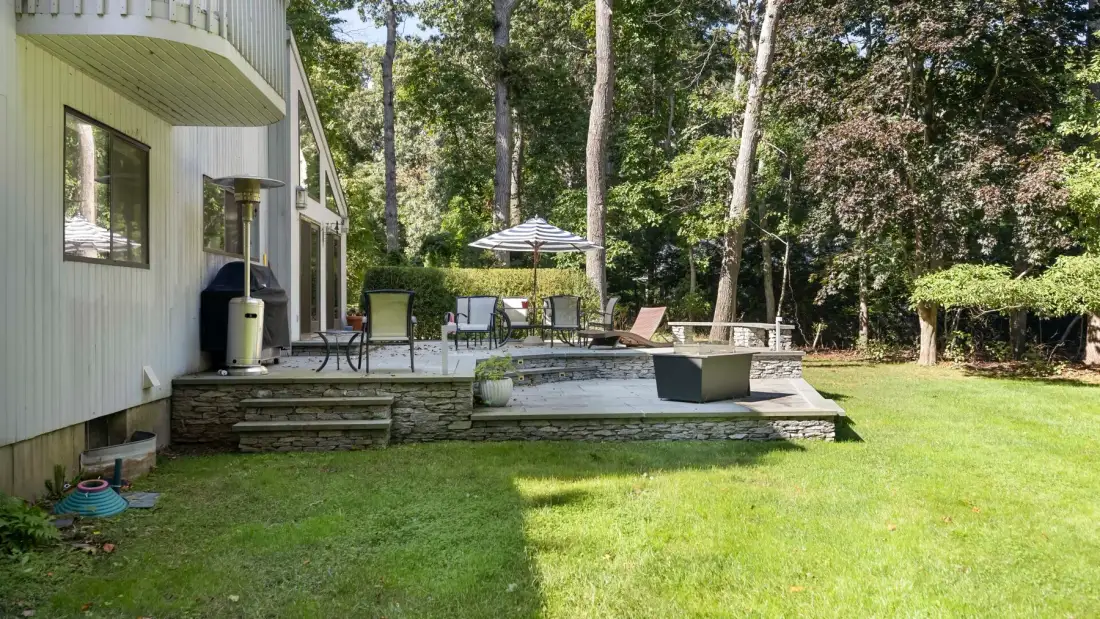 A backyard patio with outdoor seating and a striped umbrella beside the lawn.