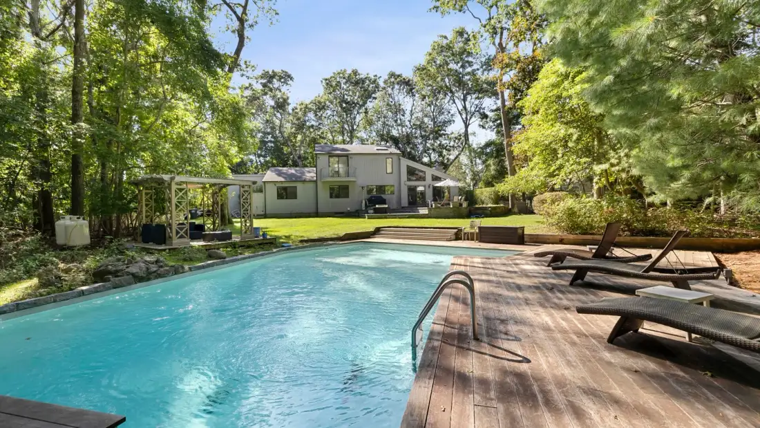 Outdoor swimming pool beside a wooden deck with lounge seating and a house beyond.