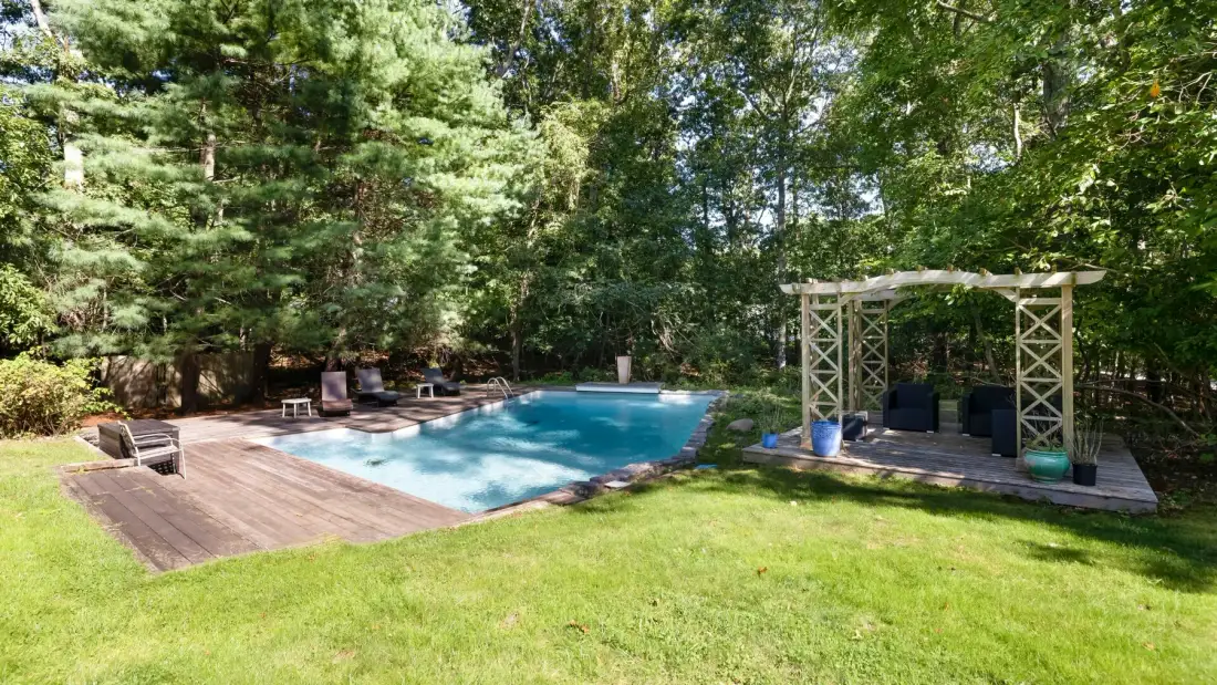 An outdoor swimming pool sits beside a wooden deck and pergola in a leafy yard.