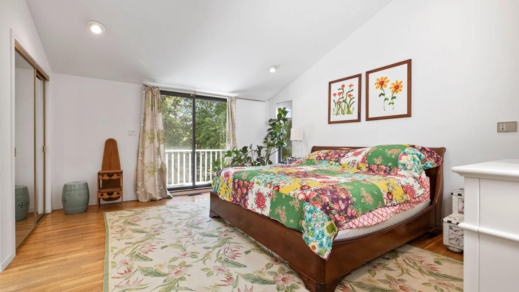 A bedroom with a colorful floral bedspread, large window, and leafy plant.