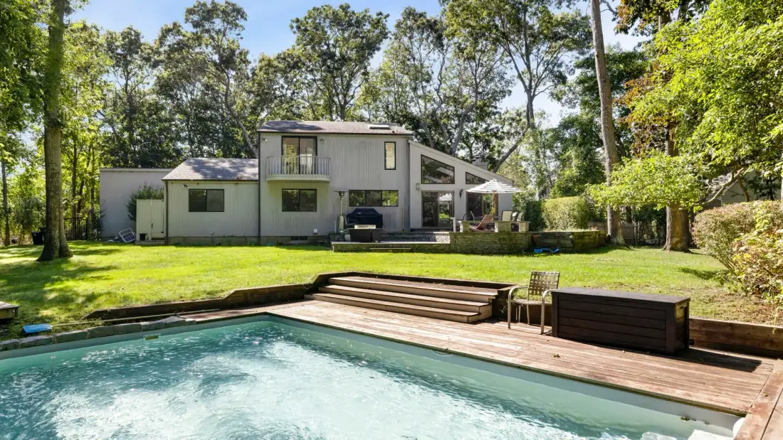 A swimming pool and wooden deck sit in front of a modern house and leafy yard.