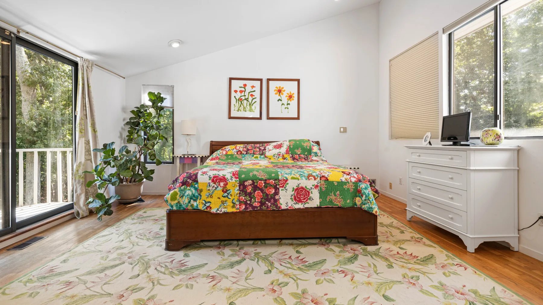A bright bedroom with a colorful floral bedspread and large windows.