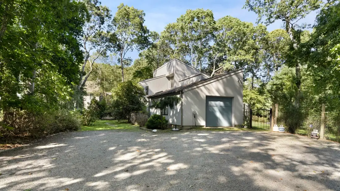 A light-colored building stands beside a gravel driveway beneath leafy trees.