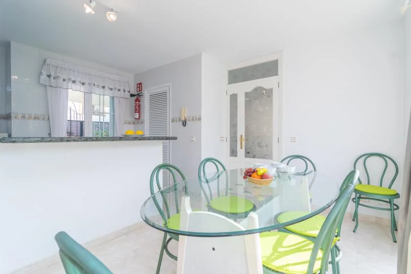 Un comedor luminoso con una mesa de cristal y asientos verdes junto a una barra de cocina.
