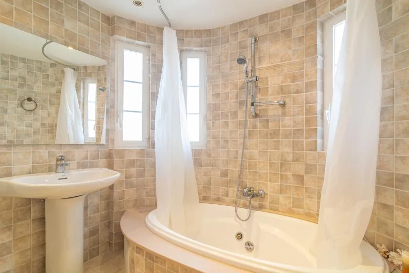 Baño con azulejos, bañera, grifería de ducha y cortinas blancas.