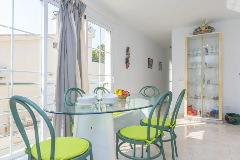 Un comedor luminoso con una mesa de vidrio, asientos verdes y grandes ventanas.