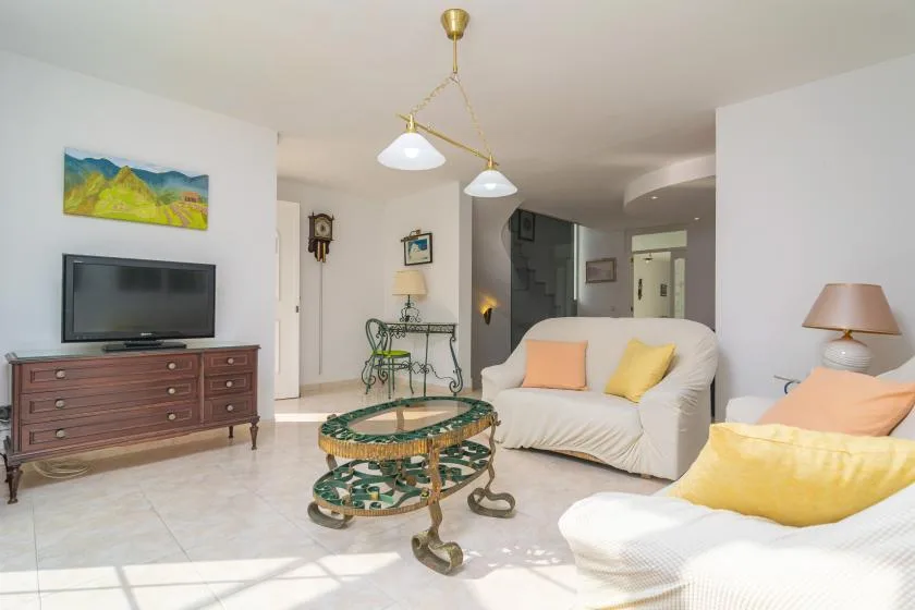 Sala de estar con asientos blancos, un televisor y una mesa de centro de vidrio.
