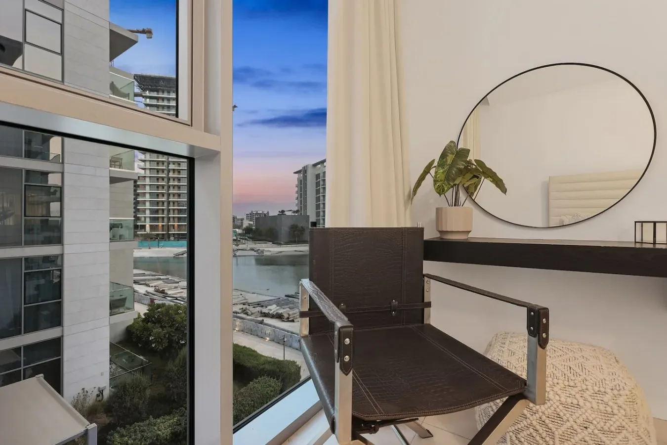 Zona de estar en un dormitorio con una silla, una planta y una gran ventana con vistas a edificios cercanos.