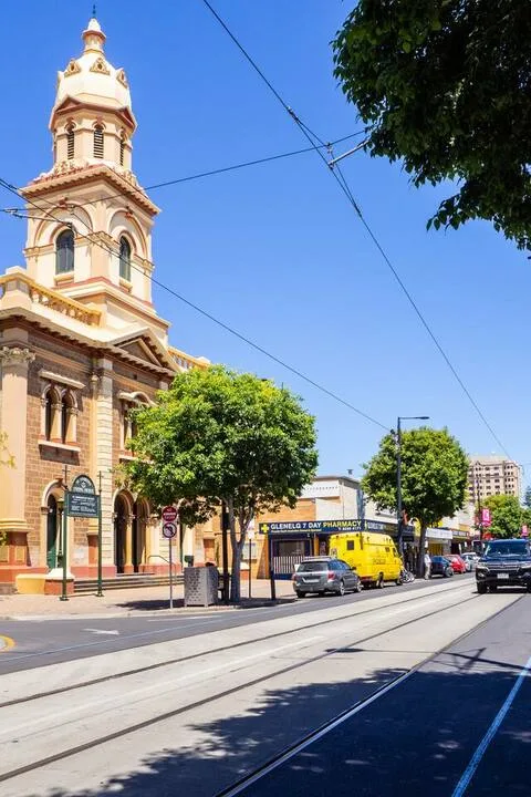 Una calle urbana con vías de tranvía pasa junto a una iglesia ornamentada y árboles frondosos.