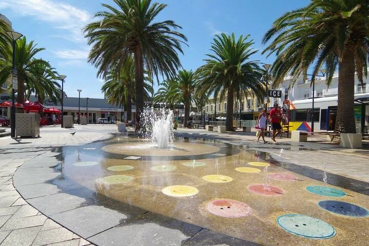 Una plaza pública bordeada de palmeras tiene una fuente circular a ras de suelo con chorros de agua.