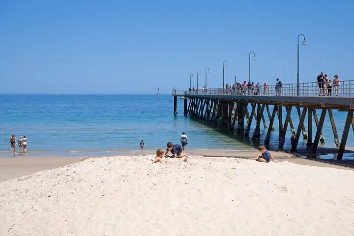 Una playa de arena se une al agua azul junto a un muelle con gente caminando.
