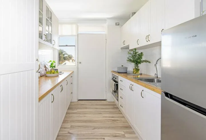 Una cocina blanca y luminosa, con encimeras a ambos lados y una ventana al fondo.