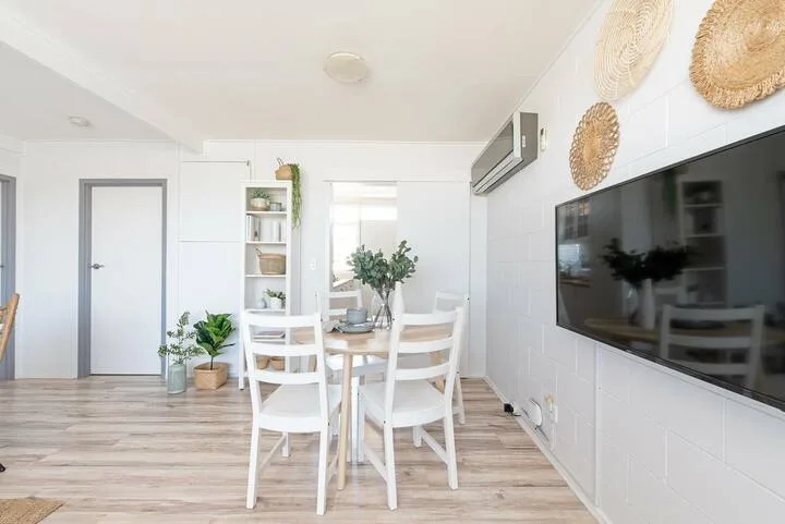 Un comedor luminoso con una mesa y sillas blancas junto a un televisor montado en la pared.