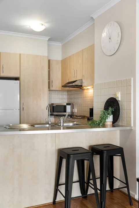 Una cocina compacta con armarios claros, un refrigerador y dos taburetes negros junto a la barra.