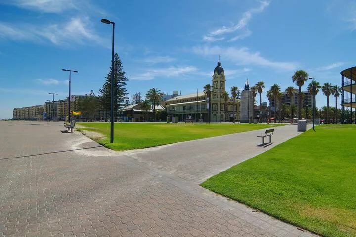 Un camino pavimentado cruza una plaza con césped frente a un edificio con torre de reloj.