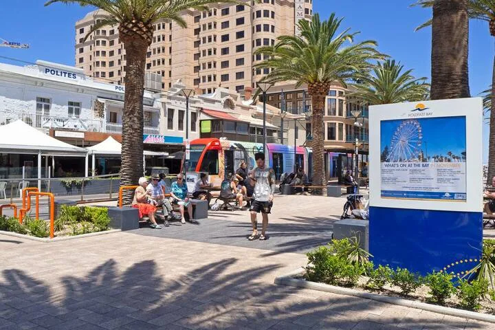 Una plaza urbana soleada con palmeras, peatones y un tranvía colorido.