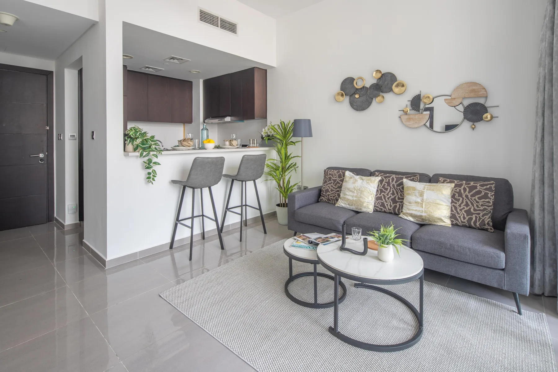 Una sala de estar con un sofá gris, dos taburetes junto a una barra de cocina y mesas de centro redondas.