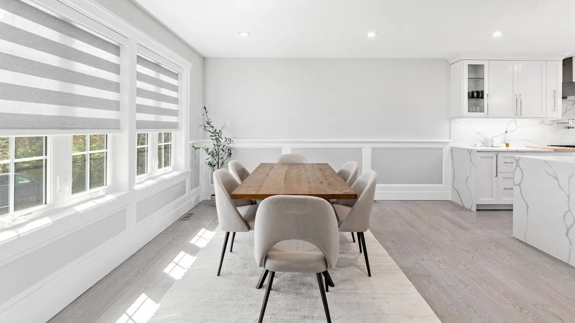 Un comedor luminoso con mesa de madera y asientos tapizados junto a grandes ventanas.