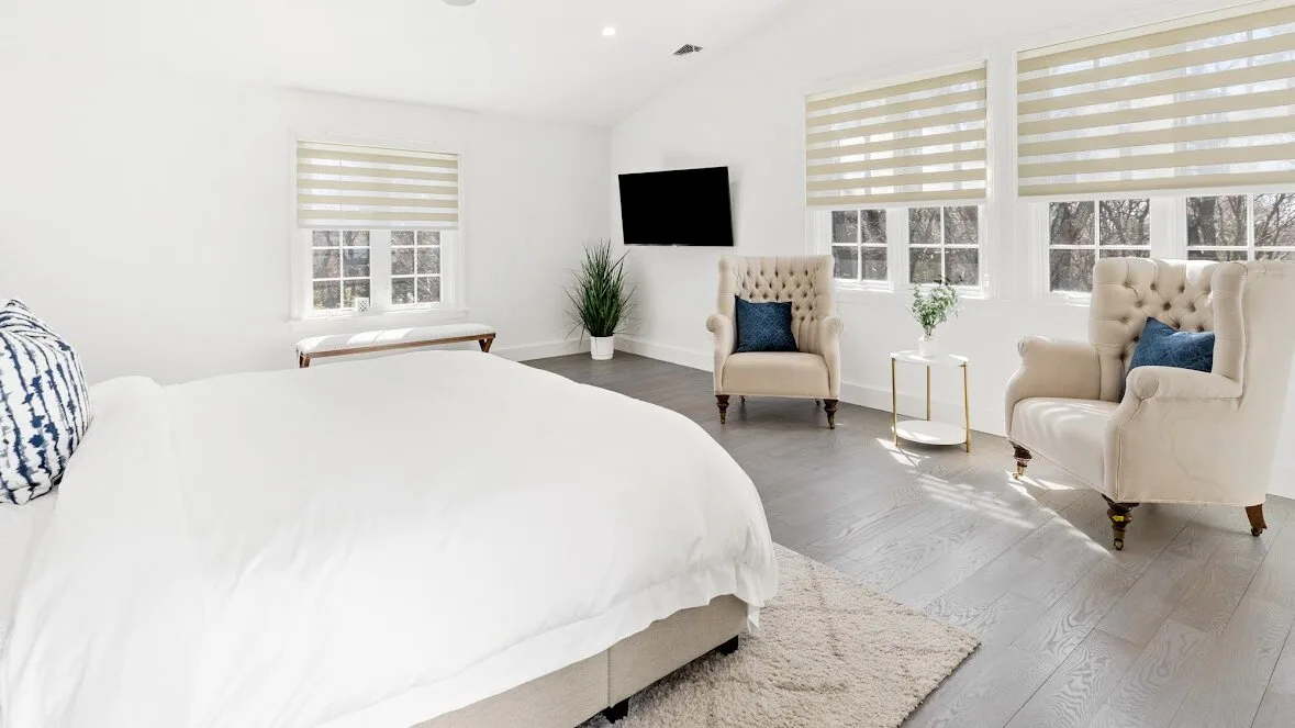 Un dormitorio luminoso con una cama grande, dos sillones y ventanas con persianas a rayas.