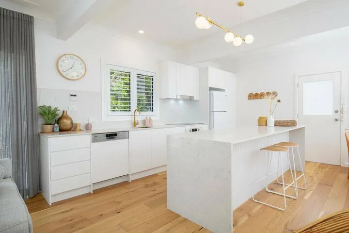 Una cocina blanca y luminosa con una isla central y suelo de madera.