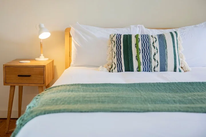 Una cama con ropa de cama blanca y una manta verde junto a una mesita de madera.