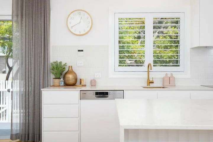 Una cocina blanca y luminosa con un grifo dorado bajo una ventana con persianas.