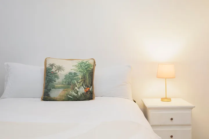 Un dormitorio con una cama blanca, un cojín estampado y una lámpara encendida.
