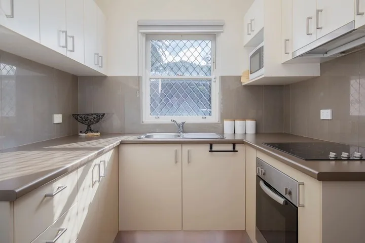 Una cocina luminosa con gabinetes claros, un fregadero bajo la ventana y una placa oscura.