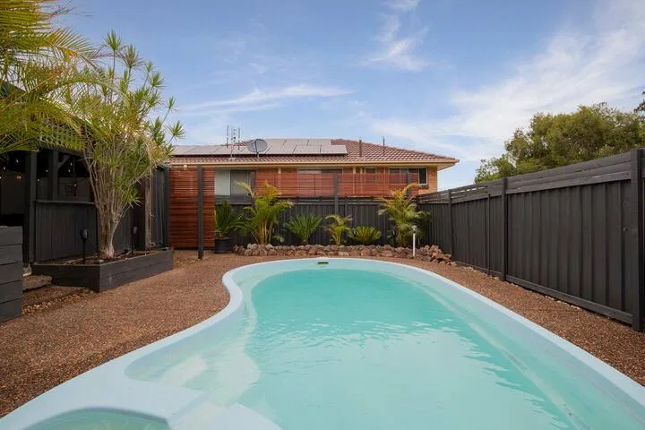 Una piscina exterior curva está junto a un patio ajardinado y cercado.